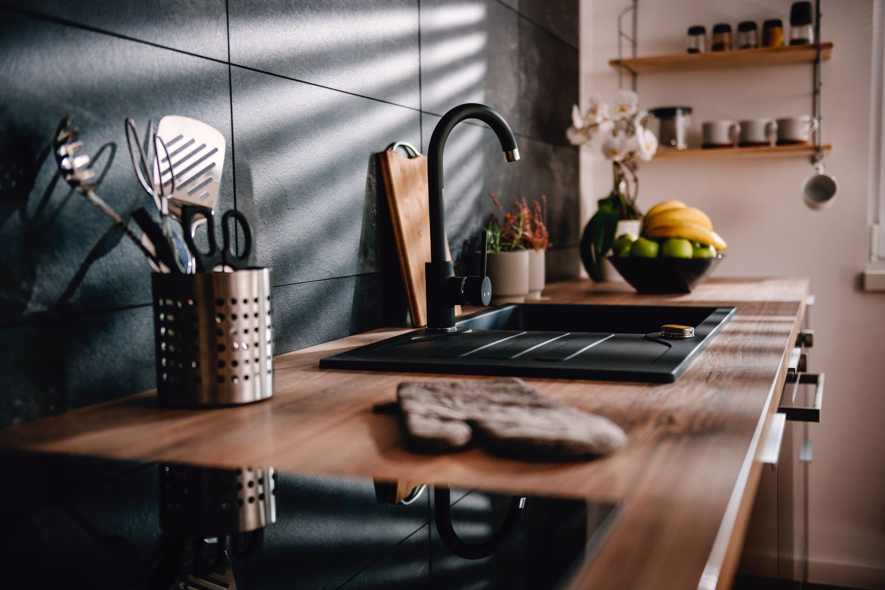 A modern kitchen countertop, featuring a black faucet, utensils, a cutting board, and a fruit bowl sleek