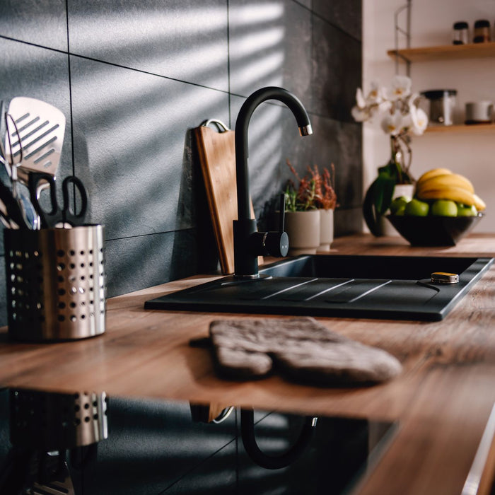 A modern kitchen countertop, featuring a black faucet, utensils, a cutting board, and a fruit bowl sleek